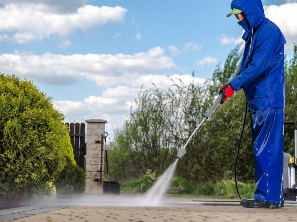 Worker in rain gear pressure washing driveway near bushes.