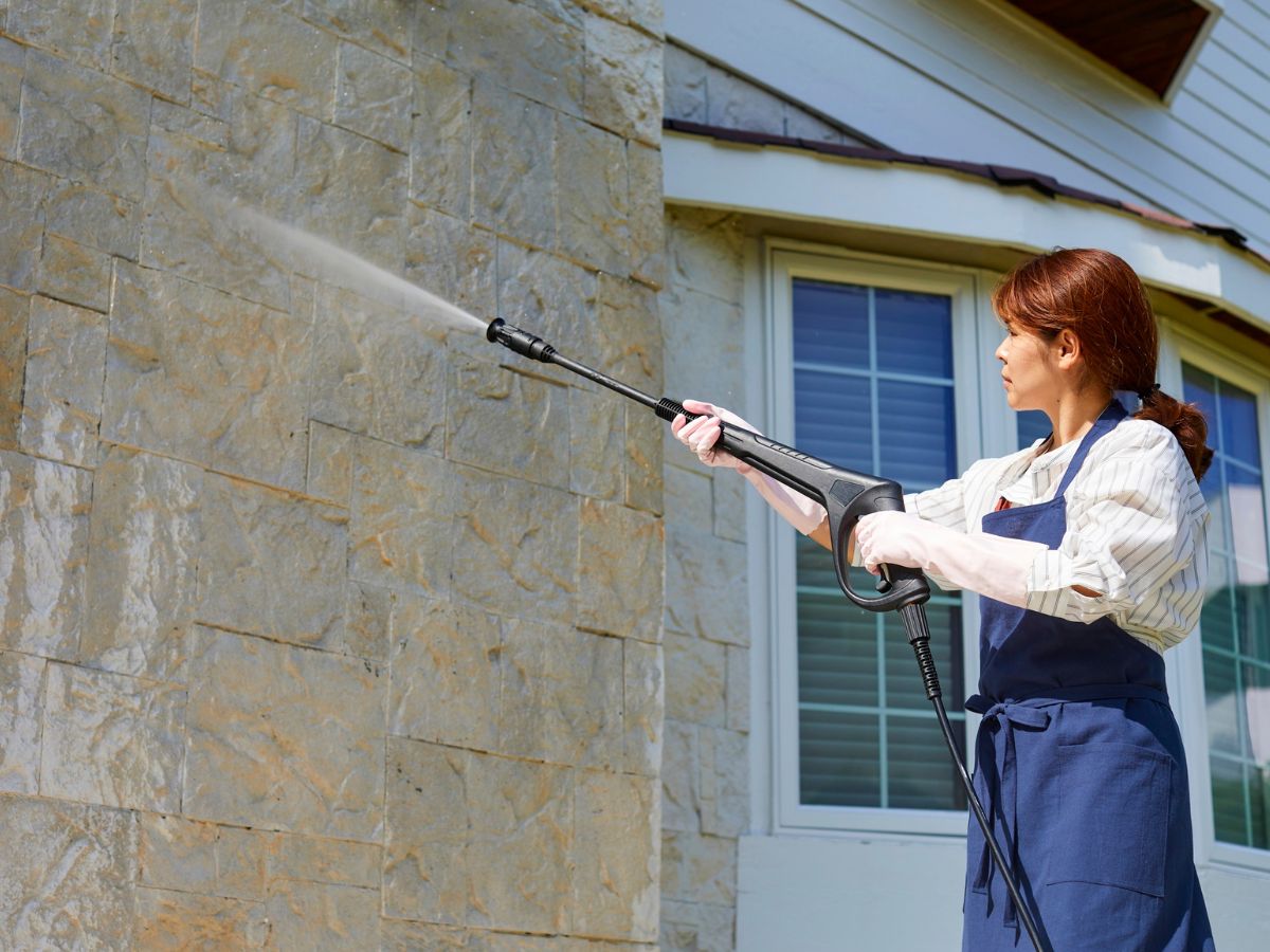 Woman pressure washing exterior stone wall of a house.